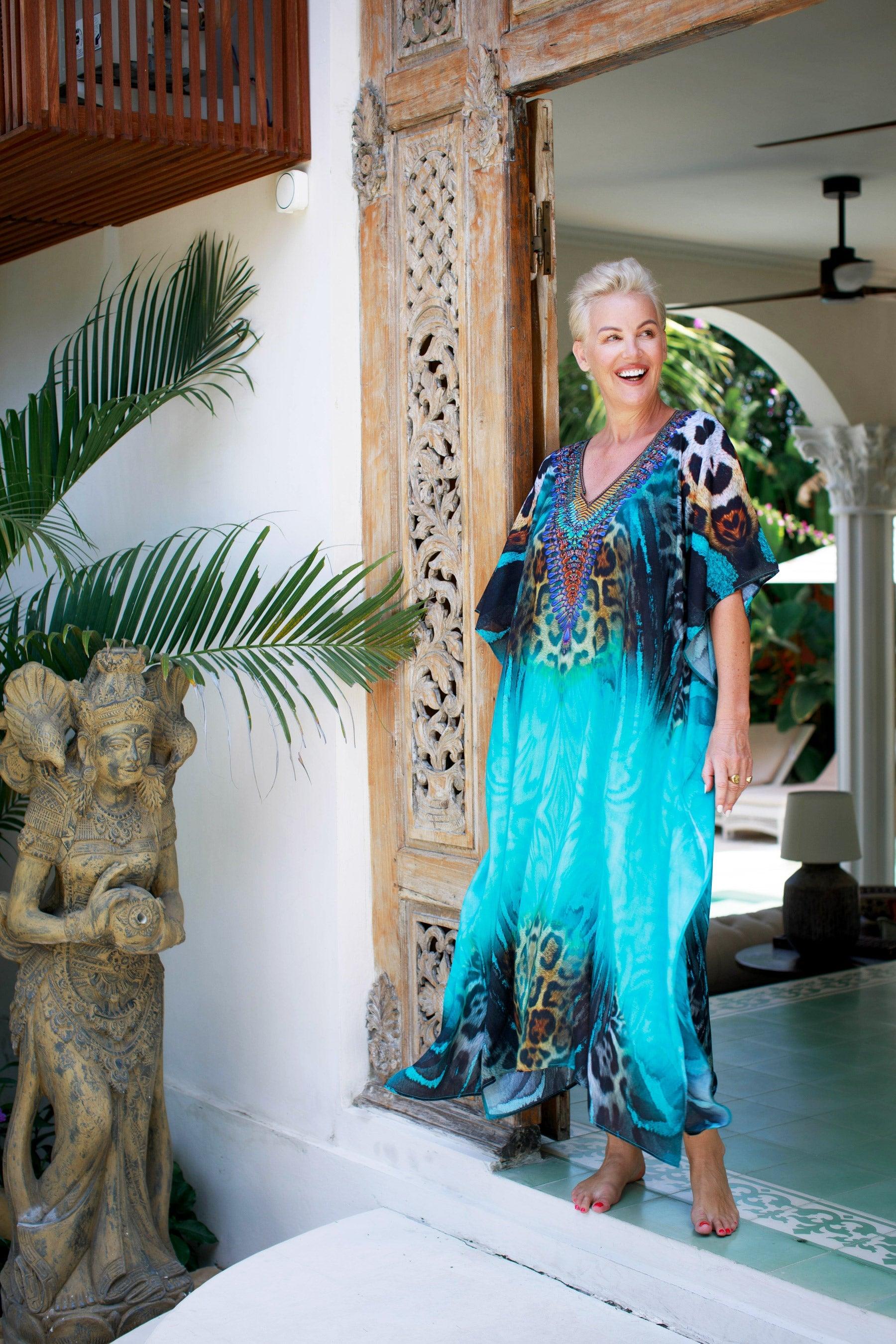 Smiling woman in a vibrant blue kaftan by Claire Powell stands in a tropical, elegant doorway.