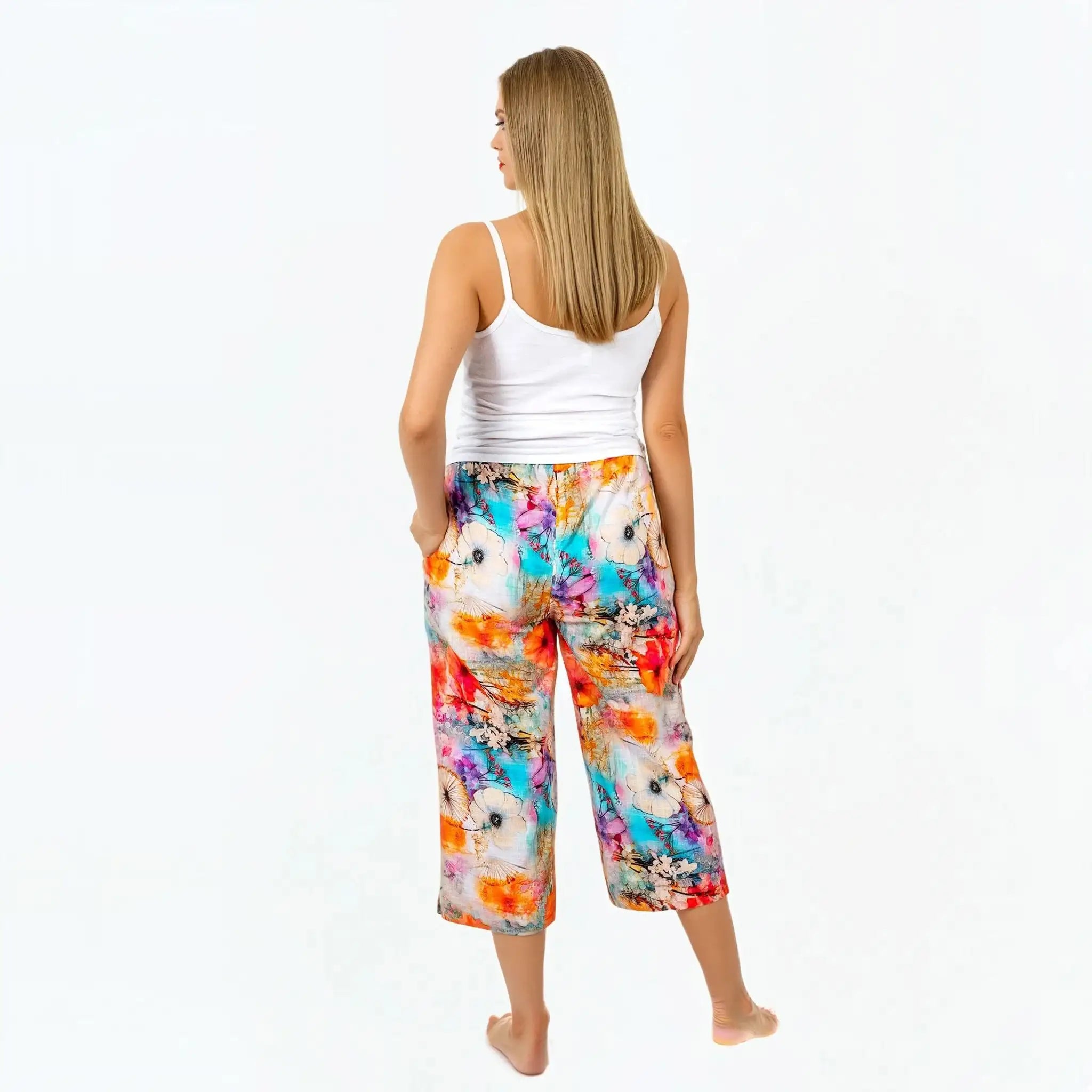 Woman in white tank top and colorful floral print Claire Powell pants, standing barefoot, back view