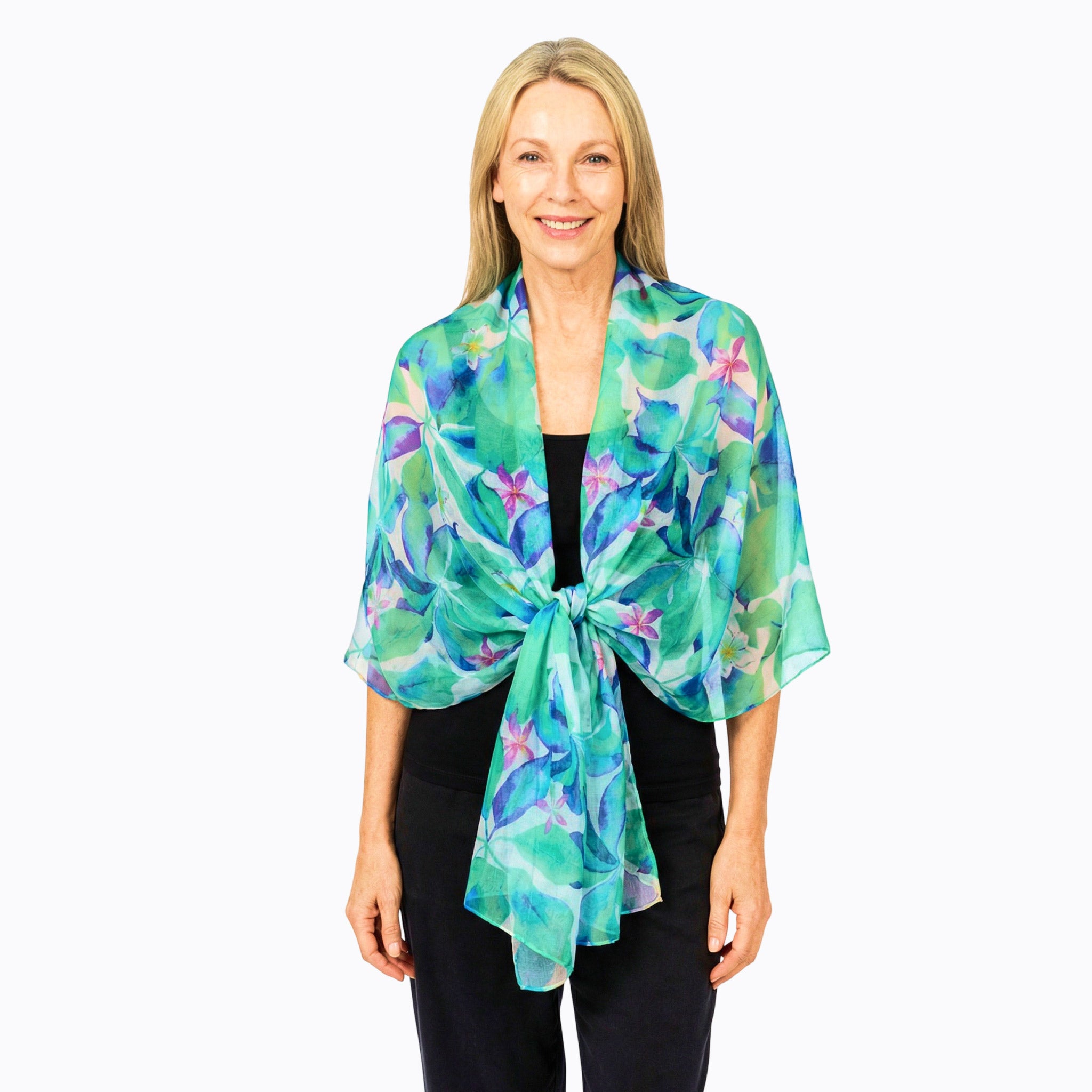 A woman modeling a lightweight, open-front shawl wrap tied at the waist. The wrap features a vibrant watercolor floral pattern in shades of teal, green, blue, and purple.