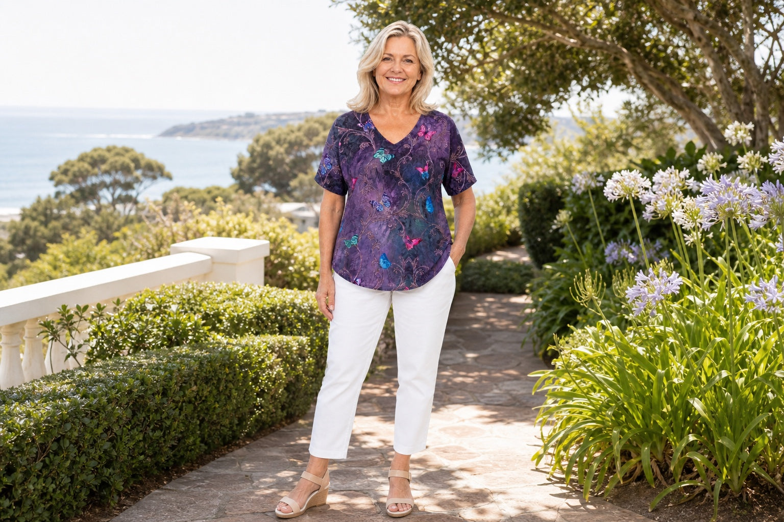 Mature woman wearing the Enchanted V-neck top with white pants in a coastal garden