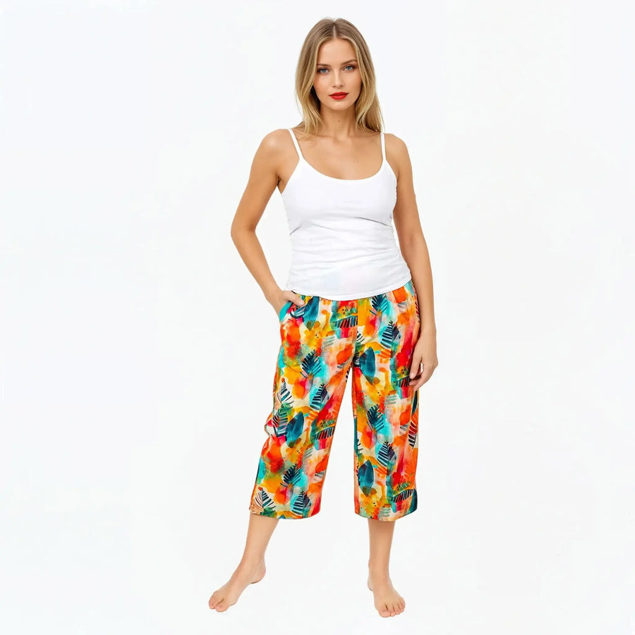 Woman wearing Claire Powell colorful tropical print pants and white tank top, barefoot on white background
