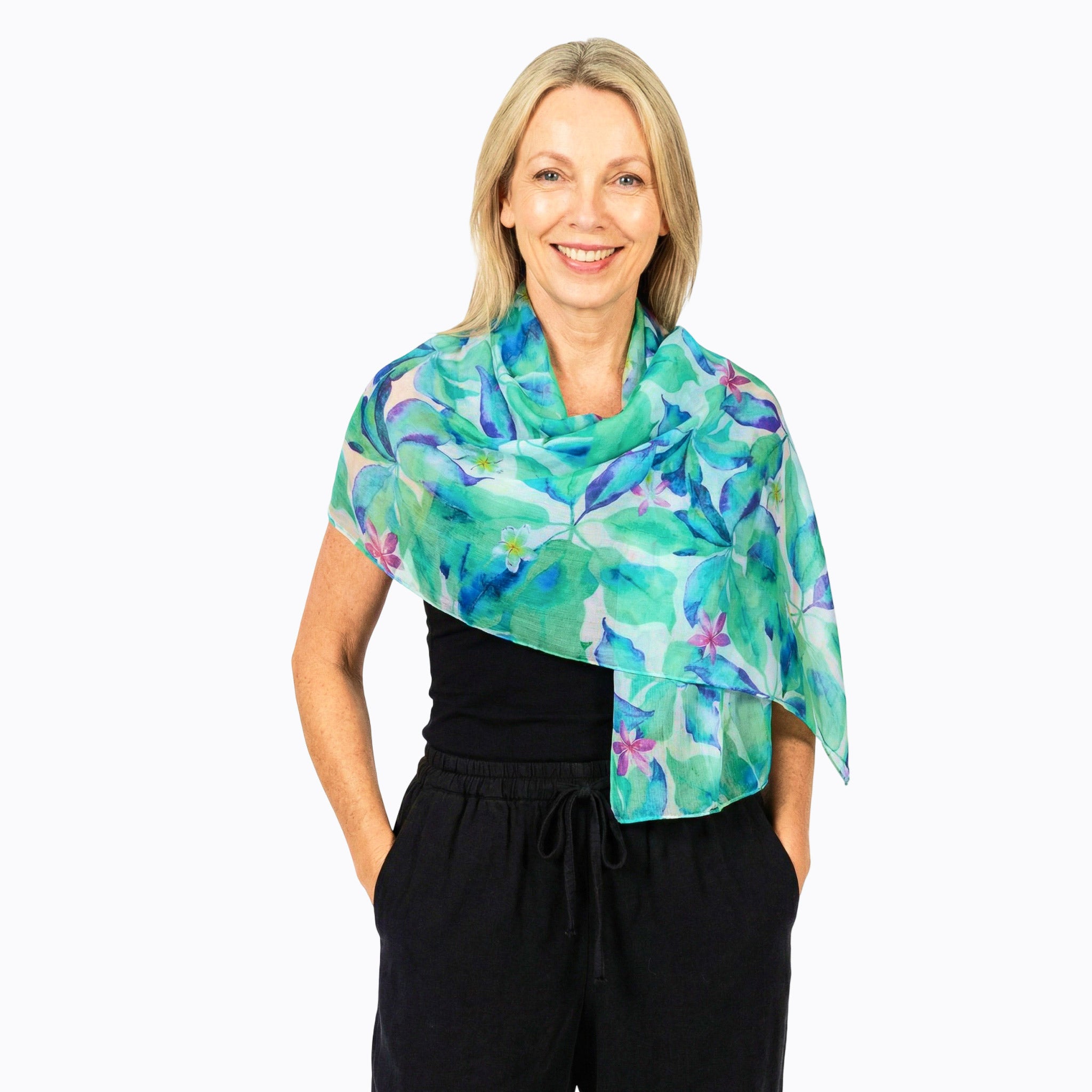 A smiling blonde woman wearing a vibrant tropical floral print shawl draped over her shoulders, paired with a black top and black drawstring pants, against a plain white background.