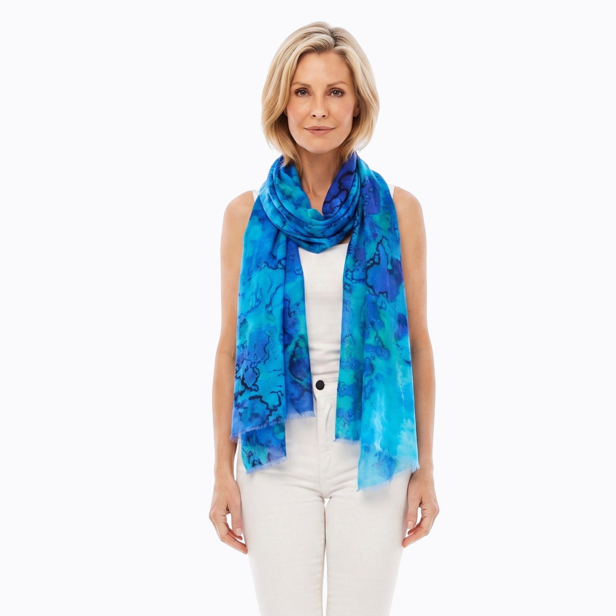 A woman modeling a lightweight blue and turquoise watercolor-patterned scarf, paired with a white sleeveless top and white pants, against a solid white background.