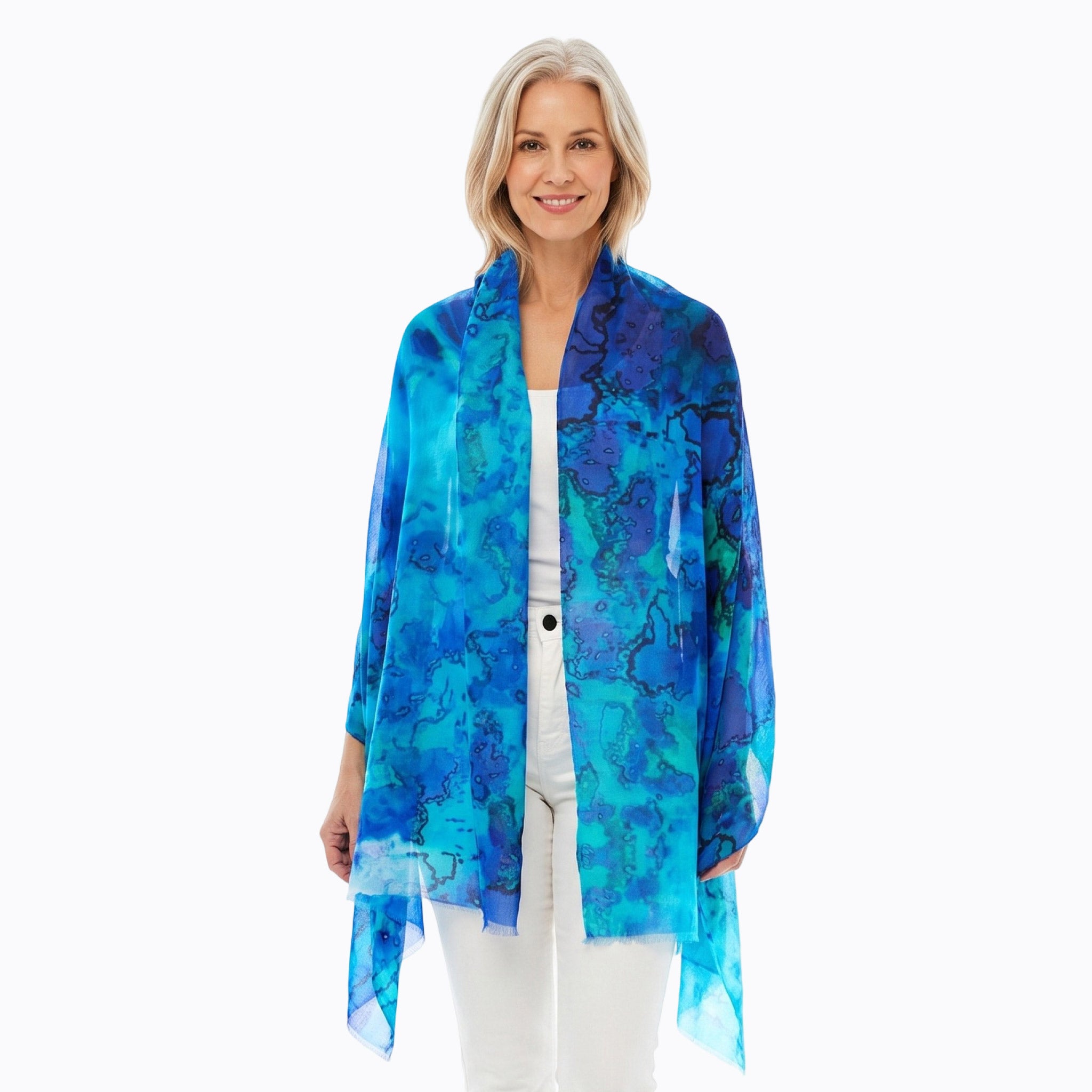 A woman modeling a lightweight, open-front shawl with an abstract watercolor pattern in shades of blue, teal, and purple, worn over a white top and white pants against a plain white background.