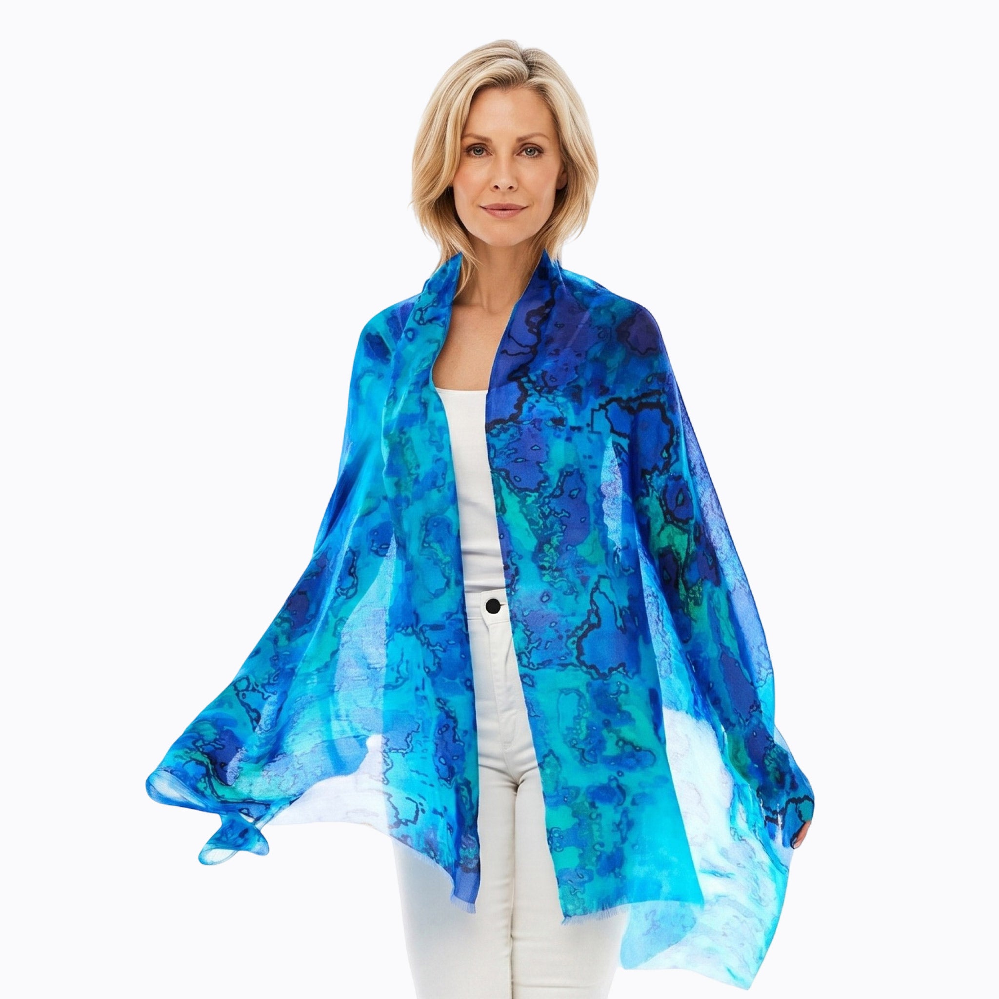 A woman modeling a lightweight sheer blue and teal abstract-patterned shawl draped over her shoulders, paired with a white top and white pants, against a plain white background.